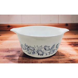 May include: White ceramic mixing bowl with a blue floral pattern. The bowl has two handles and a wide rim. The floral design features stylized flowers and leaves. The bowl is set on a wooden surface.