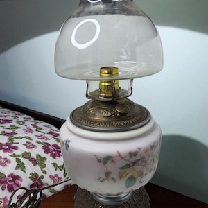 May include: Vintage oil lamp with a clear glass shade and a white base decorated with floral patterns. The lamp has a brass-colored metal base and accents. The lamp is plugged in and ready to use.