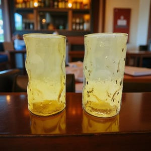 May include: Two textured, yellow-tinted glass tumblers. The glasses have an irregular, organic shape and are set on a dark wooden surface. The background is a blurred interior with bar shelving and lighting.