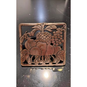 Peut inclure: Un dessous-de-plat carré de couleur cuivrée avec un motif en relief détaillé de divers fruits. Le motif comprend un ananas, des raisins et d'autres fruits, le tout dans un cadre rectangulaire. Le dessous-de-plat est probablement utilisé pour protéger les surfaces des objets chauds.