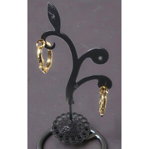 May include: A pair of gold-colored hoop earrings displayed on a black decorative earring stand. The earrings have a textured surface and are suspended from the stand's curved arms. The stand has a decorative base.