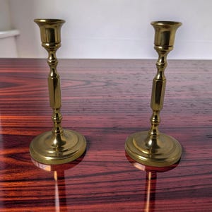 May include: A pair of polished brass candlesticks featuring a flared top, a slender stem with a central hexagonal section, and a wide circular base. The metallic surface reflects light, highlighting the smooth, reflective finish of the metal.