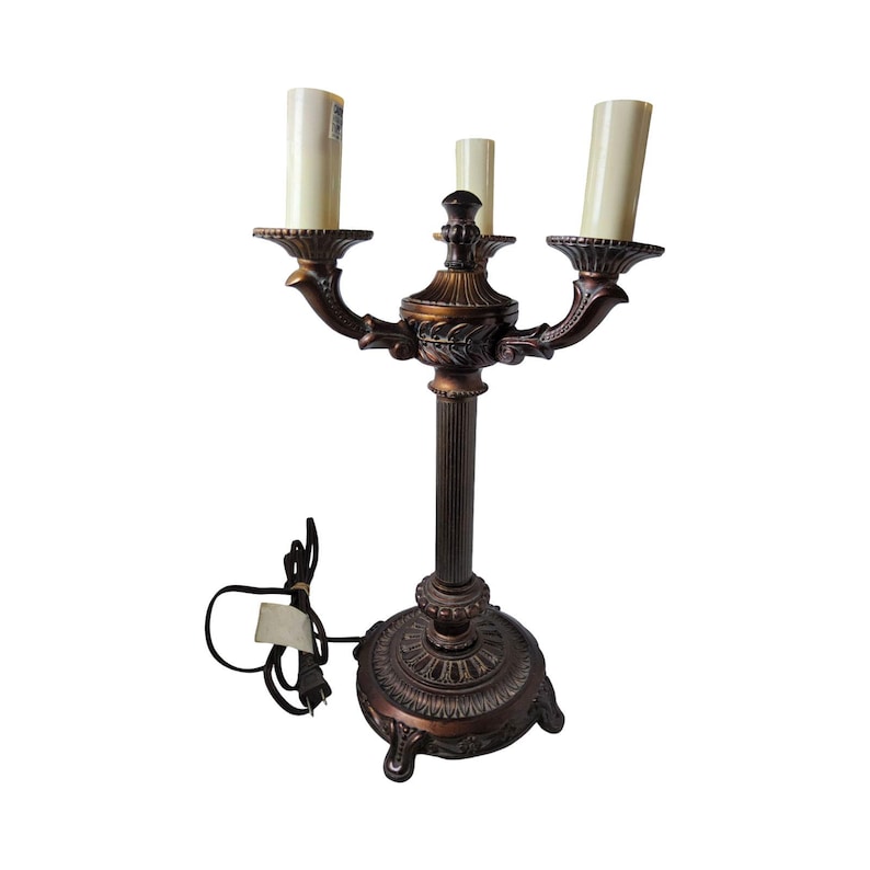 May include: A three-light candelabra lamp with a dark bronze-toned finish, featuring a fluted central column, an ornate circular base with three feet, and three cream-colored cylindrical light covers extending from decorative arms.