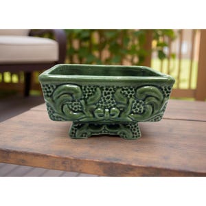 Op de afbeelding: Een groene keramische plantenbak met een rechthoekige vorm en sierlijke bloemendetails. De plantenbak staat op kleine voetjes en heeft een glanzende afwerking. Ideaal voor kleine planten of vetplanten.
