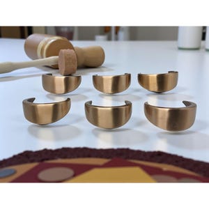 Könnte beinhalten: Sechs halbkreisförmige Metallobjekte in Bronzefarbe sind auf einer weißen Oberfläche angeordnet. Ein Holzhammer mit hellem braunen Kopf und Griff liegt im Hintergrund. Die Objekte scheinen Beschläge oder dekorative Elemente zu sein.