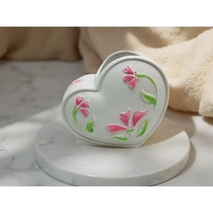 May include: A white heart-shaped ceramic vase with pink floral designs and green stems and leaves. The vase is sitting on a white marble surface.