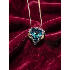 Puede incluir: Un collar con colgante en forma de corazón con una cadena plateada. El colgante presenta una gran piedra preciosa azul-verdosa rodeada de pequeños cristales transparentes y detalles metálicos dorados. El collar descansa sobre una superficie de terciopelo rojo oscuro.