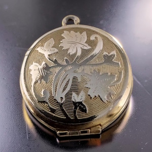 May include: A gold-toned, round locket with a floral design. The locket features an engraved pattern of leaves and vines in a lighter gold color. It has a small loop at the top for a chain and a hinged opening at the bottom.