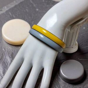May include: Two smooth, circular bangle bracelets worn on a white mannequin hand. One bangle is a solid, glossy mustard yellow, and the other is a solid, glossy slate gray. Both bracelets feature a simple, rounded profile with a polished finish.