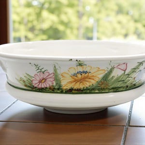 May include: A white ceramic bowl featuring a hand-painted floral design around the exterior. The pattern includes yellow and pink flowers with green foliage and stems, accented by a thin green line encircling the base of the bowl's rim.