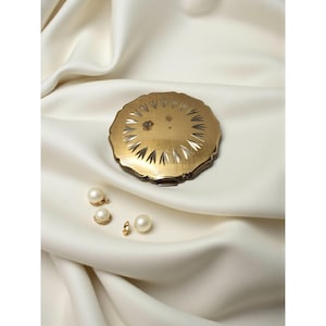 May include: A gold-toned compact with a sunburst design and a small gemstone is displayed on a white satin surface. Three pearl earrings and a single pearl stud are also present, creating an elegant, vintage aesthetic.