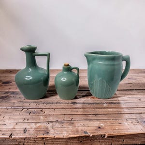 May include: Three teal ceramic vessels of varying sizes sit on a wooden surface. The collection includes a pitcher, a small jug with a handle and cork, and a vase with a handle. The pitcher has a decorative floral design.