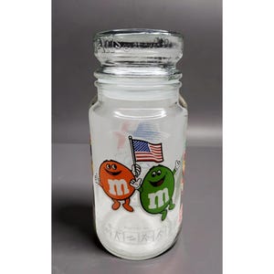 May include: Clear glass jar with a silver lid. The jar features cartoon characters holding an American flag. The characters are orange and green with the letter "m" on their bodies. The jar has a wide mouth and a cylindrical shape.
