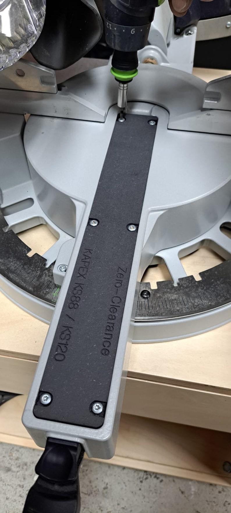 May include: A gray and black zero-clearance insert for a Kapex KS88 / KS120 saw. The insert has a black rubber strip and is being secured with a screw.
