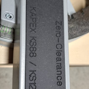 May include: Close-up of a black metal plate with the text "KAPEX KS88 / KS120 Zero-Clearance" engraved on it. The plate is attached to a silver machine with screws.