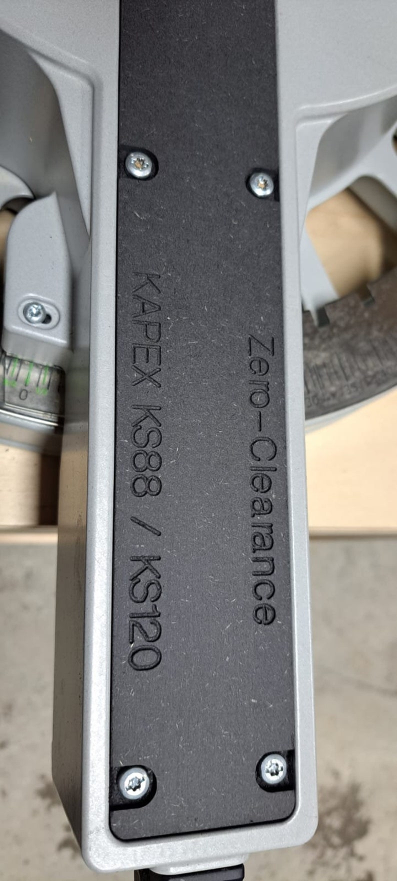 May include: Close-up of a black metal plate with the text "KAPEX KS88 / KS120 Zero-Clearance" engraved on it. The plate is attached to a silver machine with screws.