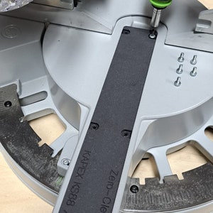 May include: A black and silver power tool with a green handle is being used to tighten a screw on a gray and black saw. The saw has a black label that reads "Zero-Clearance KAPEX KS88/ KS120".