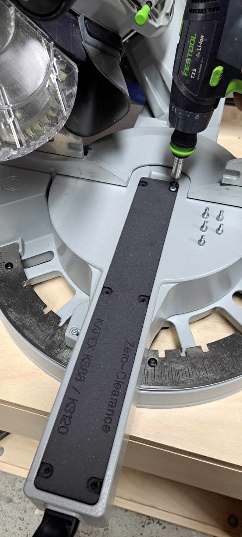 May include: A black and silver power tool with a green handle is being used to tighten a screw on a gray and black saw. The saw has a black label that reads "Zero-Clearance KAPEX KS88/ KS120".