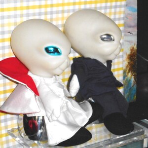 May include: Two alien figurines with large, smooth, light-colored heads and reflective eyes. One figure wears a white outfit with a red-lined cape, while the other wears a dark blue suit. Both have black shoes and are positioned side by side.