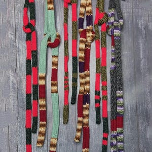 May include: Six long, thin, hand-knitted scarves in various color combinations including red, dark green, mint green, brown, purple, and variegated yarn. The scarves are draped over a curved wooden hanger, each featuring distinct striped patterns.