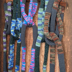 May include: A collection of five long, knitted scarves featuring a ribbed texture. Each scarf combines solid dark gray sections with variegated yarn segments in vibrant colorways including purple, blue, pink, green, and earthy brown tones.