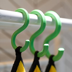 May include: Three vibrant green S-shaped hooks suspended from a white rail. The hooks support black and yellow towels. The hooks are made of a smooth material, likely plastic, and are designed for hanging items.