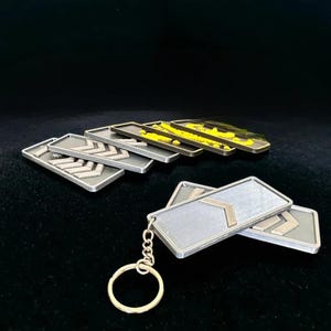 Puede incluir: Llaveros rectangulares plateados con diseños de galones. Algunos llaveros tienen un diseño de galón dorado. Un llavero está unido a un llavero plateado. Los llaveros están dispuestos sobre una superficie oscura.