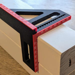 Pode incluir: Uma ferramenta de esquadro rápido preta e vermelha, apoiada em um bloco de madeira branco. A ferramenta possui uma régua com medições de até 70 mm, projetada para medições precisas de ângulos em carpintaria e construção.