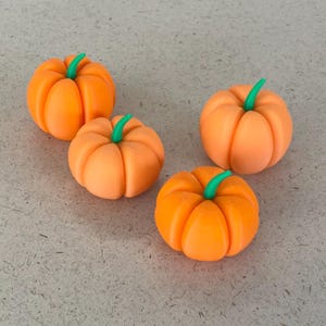 Può includere: Quattro piccole zucche arancioni con steli verdi sono disposte su una superficie strutturata grigio chiaro. Le zucche sono di un colore arancione vibrante e hanno una consistenza a coste, che conferisce loro un aspetto realistico. Gli steli verdi aggiungono un tocco di contrasto.