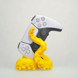 May include: A white and black video game controller is held by a bright yellow octopus tentacle base. The controller has buttons and directional pads. The octopus base has suction cups.