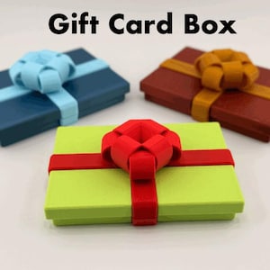 Pode incluir: Três caixas retangulares para cartões-presente em azul, vermelho e verde, cada uma adornada com uma fita e um laço a condizer. O texto "Gift Card Box" é exibido acima das caixas. As caixas estão dispostas sobre uma superfície branca.