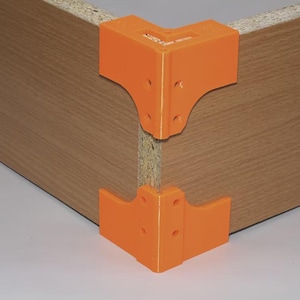 Op de afbeelding: Twee feloranje hoekklemmen zijn bevestigd aan de hoek van een houten paneel. De klemmen zijn gemaakt van plastic en hebben verschillende gaten voor schroeven. Het houten paneel heeft een lichtbruine houtnerf afwerking.