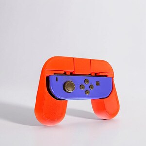 May include: An orange and purple video game controller grip. The grip is orange and surrounds a purple controller with gold and black buttons. The controller is designed to enhance the gaming experience.