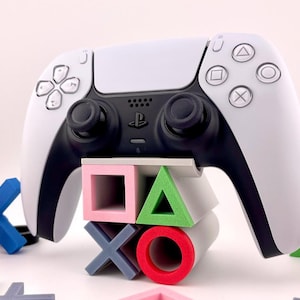 May include: A white and black PlayStation 5 controller sits on a stand with colourful geometric shapes. The stand features a pink square, a green triangle, a grey X, and a red circle. Additional shapes are scattered around.