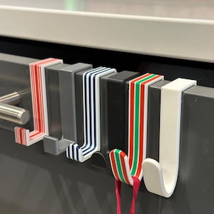May include: A row of decorative over-the-door hooks in various colors. The hooks are white, gray, and black, with striped and solid designs in red, green, and white. The hooks are designed to hang over a door or cabinet.