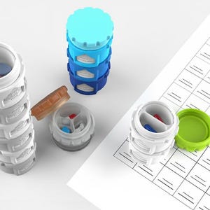 Peut inclure: Organisateurs de pilules empilables blancs, bleus et verts. Les contenants à pilules sont cylindriques avec un intérieur segmenté. Certains contenants sont ouverts, révélant des pilules colorées. Une feuille de papier avec un motif quadrillé est en arrière-plan.