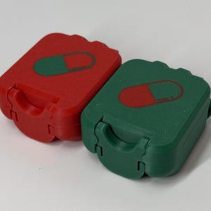 Pode incluir: Duas pequenas caixas retangulares para comprimidos. Uma é vermelha com um gráfico de comprimido verde, a outra é verde com um gráfico de comprimido vermelho. Cada caixa tem uma alça e uma tampa articulada. As caixas estão em uma superfície branca.