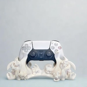 Könnte beinhalten: Ein weiß-schwarzer Gamecontroller liegt auf einem dekorativen Ständer. Der Ständer ist weiß und hat ein Design aus Totenköpfen und knochenartigen Strukturen. Der Controller hat eine dunkelgraue Mitte und weiße Seiten.