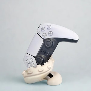 May include: A white and black video game controller on a cream-coloured octopus-shaped stand. The controller features buttons and directional pads. The stand has a curved design with suction cup details.