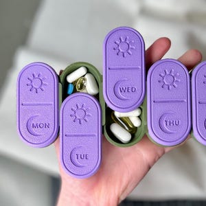 Pode incluir: Um organizador de comprimidos semanal roxo e verde com compartimentos rotulados para cada dia da semana. Os compartimentos são preenchidos com vários comprimidos e cápsulas. O organizador é segurado em uma mão.