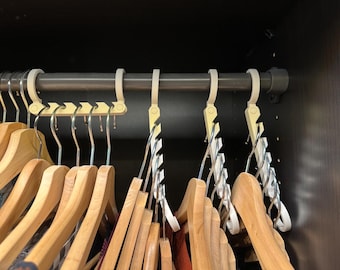 May include: A closet interior with a metal rod holding various hangers. Wooden hangers are in the foreground, while plastic space-saving hangers and a multi-hanger are visible. The closet is dark, with the hangers in the center.