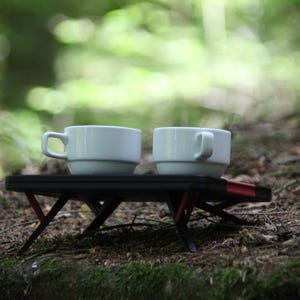 Peut inclure: Deux tasses à café blanches avec des poignées sont posées sur une petite table pliante noire avec des accents rouges. La table est installée à l'extérieur sur un lit de feuilles et de mousse, avec un arrière-plan vert flou.