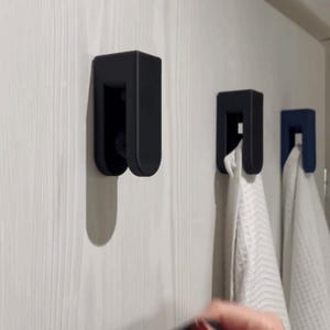 Op de afbeelding: Zwarte aan de muur gemonteerde haken met een U-vormig ontwerp. Twee haken houden witte handdoeken vast, terwijl een derde haak leeg is. De haken zijn gemonteerd op een witte wand met houten panelen. Een persoon houdt een donkerblauwe handdoek vast.