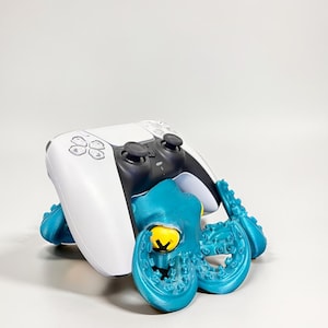 May include: A white and black PlayStation 5 controller sits on a teal octopus-shaped stand. The octopus has a yellow eye and detailed tentacles. The stand is designed to hold the controller securely.