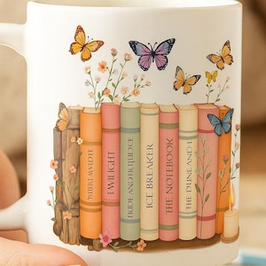 May include: White ceramic mug featuring a design of colorful butterflies and a row of books. The books have titles like "Twilight" and "The Notebook." Floral accents and a candle complete the design. A perfect gift for book lovers.