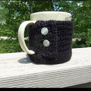 May include: A white ceramic mug with a cream-colored handle is wrapped in a dark gray crocheted cozy. The cozy is secured with two large, pearlescent buttons. The mug sits on a white wooden surface, with green foliage in the background.
