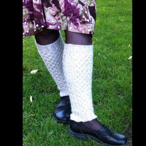 May include: A pair of light gray knitted leg warmers with a diamond pattern, worn over black tights. The leg warmers are paired with black shoes, and the background is green grass.