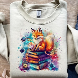 May include: A cream-colored sweatshirt featuring a vibrant illustration of a fox sitting atop a stack of books. The fox is orange and white, with blue eyes. A clock, a crystal ball, and other magical elements surround the fox.
