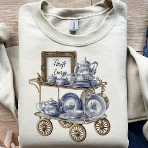 May include: Cream-colored sweatshirt featuring a detailed illustration of a vintage tea cart. The cart is laden with blue and white china, including teacups, teapots, and plates. The text "Thrift Energy" is displayed in a framed sign.