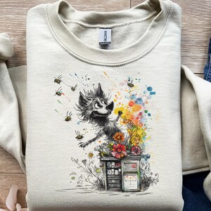 Op de afbeelding: Een lichtbeige sweatshirt met een grillige illustratie. Het ontwerp bevat een cartoonwolf met bijen, bloemen en een kleurrijke splash. De sweatshirt heeft een ronde hals en lange mouwen.